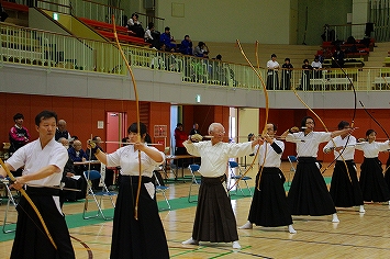 弓道大会で選手が並んで弓を構えているところ。山本会長もまだまだ現役です。